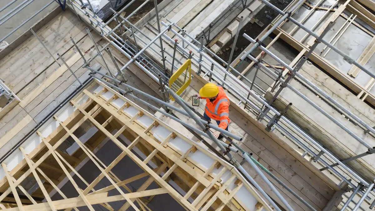Construction worker on building site showing housebuilding, roof trusses, scaffolding and completed houses. Cornwall, UK