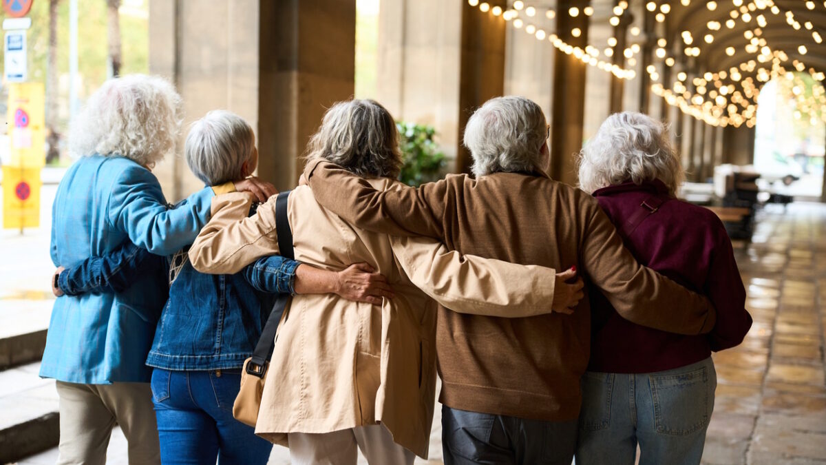 Five senior women walking arm in arm down a city archway, joyful and supportive, celebrating friendship, health and togetherness