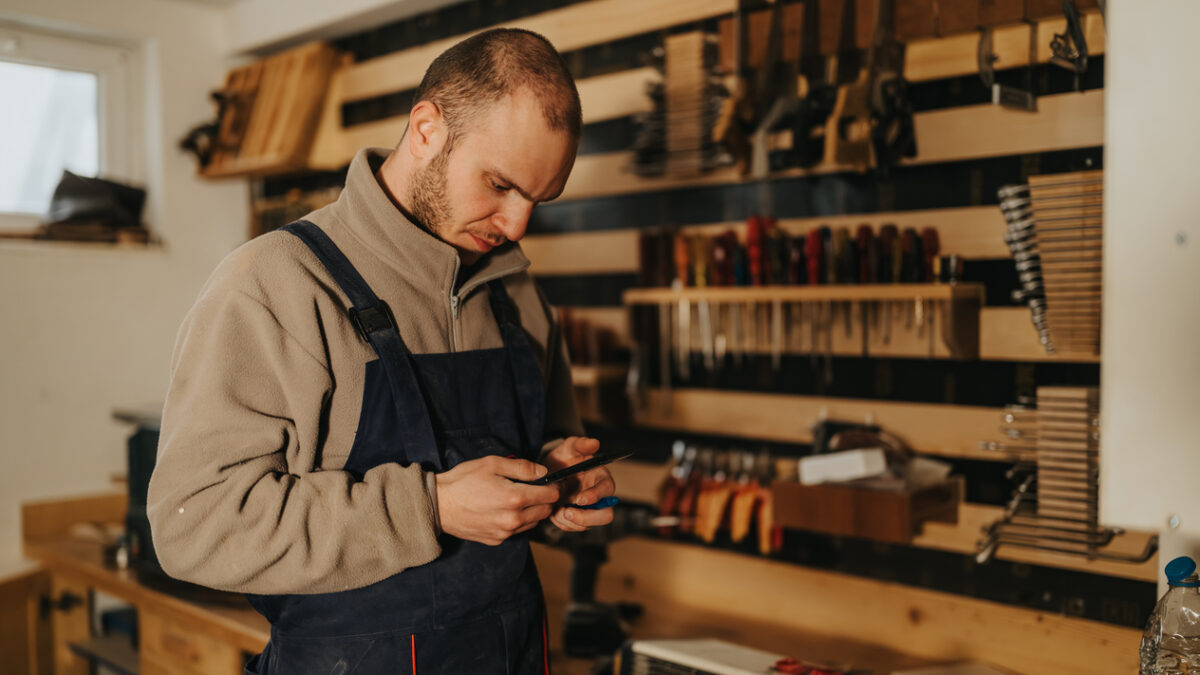 A male woodworker wearing overalls examines his phone while standing at a busy workshop bench. He checks references and measurements surrounded by hand tools, clamps and wood materials.
