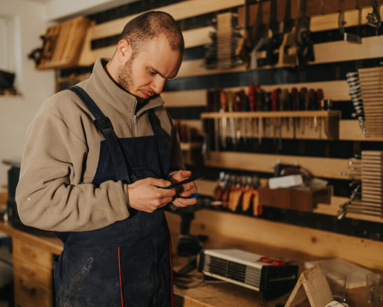 A male woodworker wearing overalls examines his phone while standing at a busy workshop bench. He checks references and measurements surrounded by hand tools, clamps and wood materials.