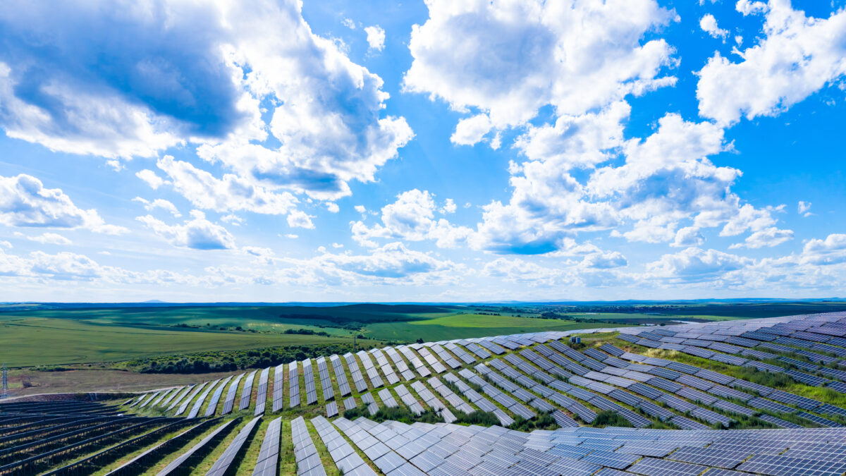 Solar panels stretch across a large area in the countryside. The sun shines bright on this field of panels with green land and blue sky in view.