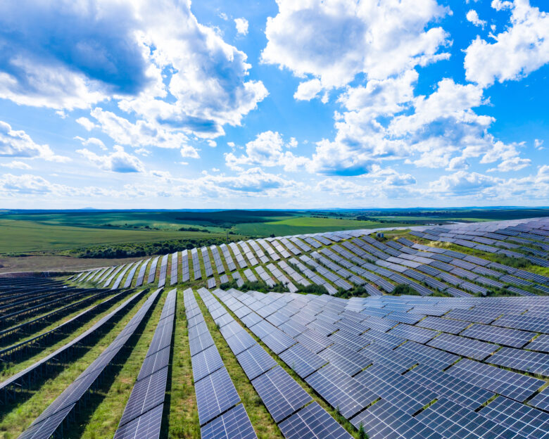 Solar panels stretch across a large area in the countryside. The sun shines bright on this field of panels with green land and blue sky in view.
