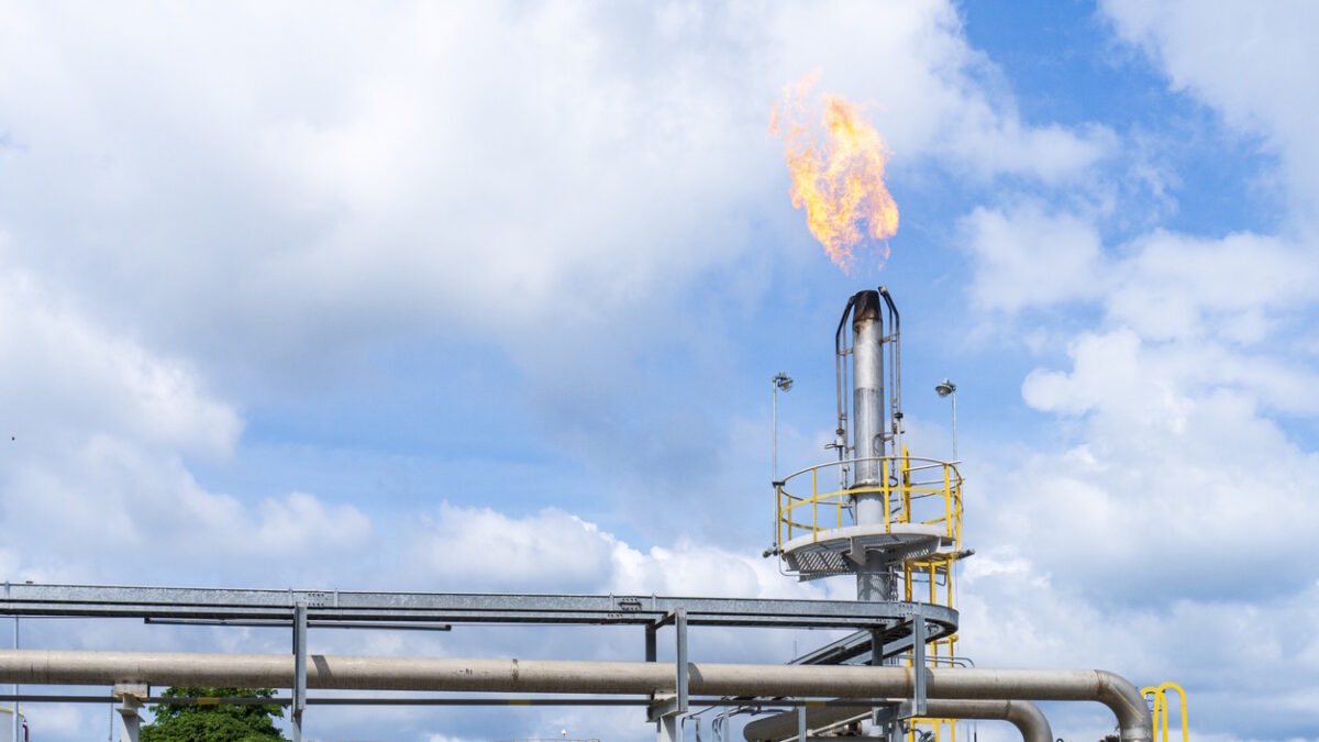 Large gas flare burning dramatically from the top of a flare stack against a blue sky, with foreground pipelines, red storage tanks, and yellow railings at an industrial plant facility.