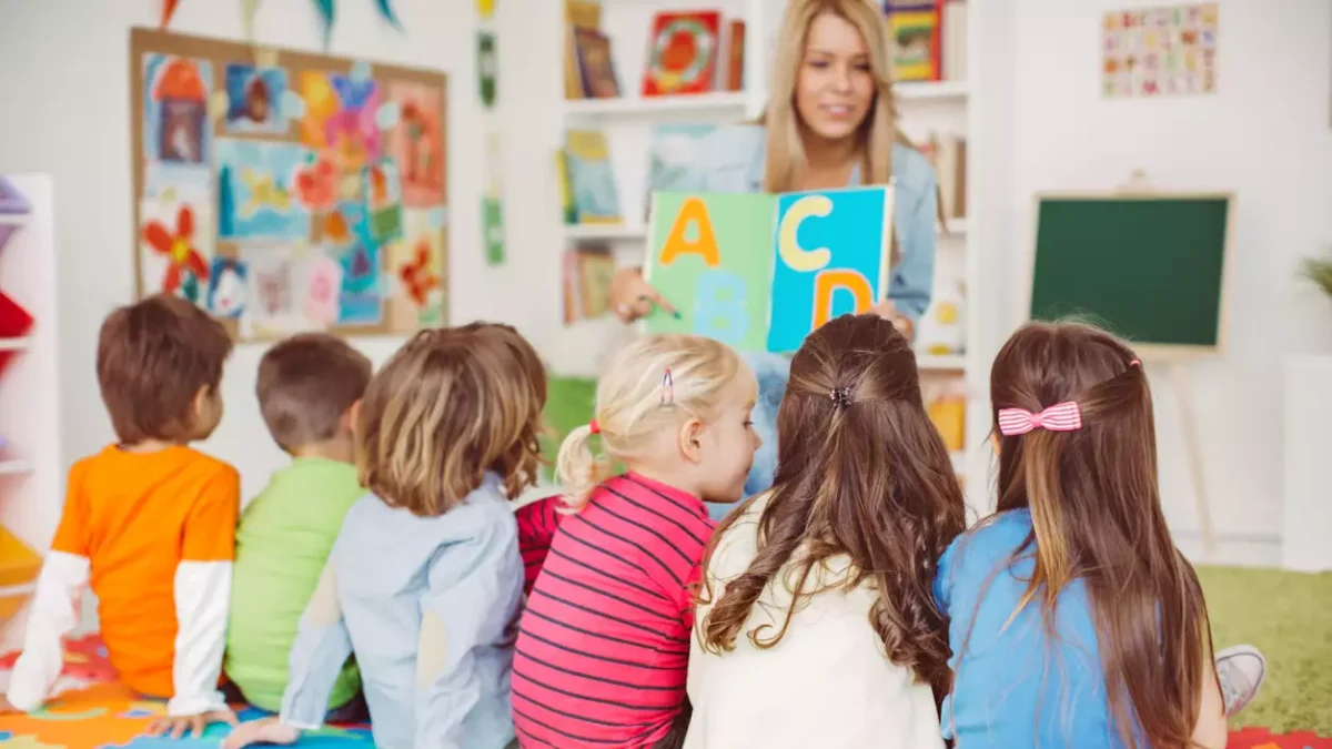 Teacher with a group of preschool children in a nursery. The children are sitting on the floor and listening teacher. Learning letters. In the background we can see a shelf with some, toys, black board and books. View from behind.