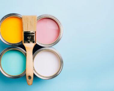 Brush with wooden handle on open cans on blue pastel background. Yellow, white, pink, turquoise colors. Renovation concept. Place for text.