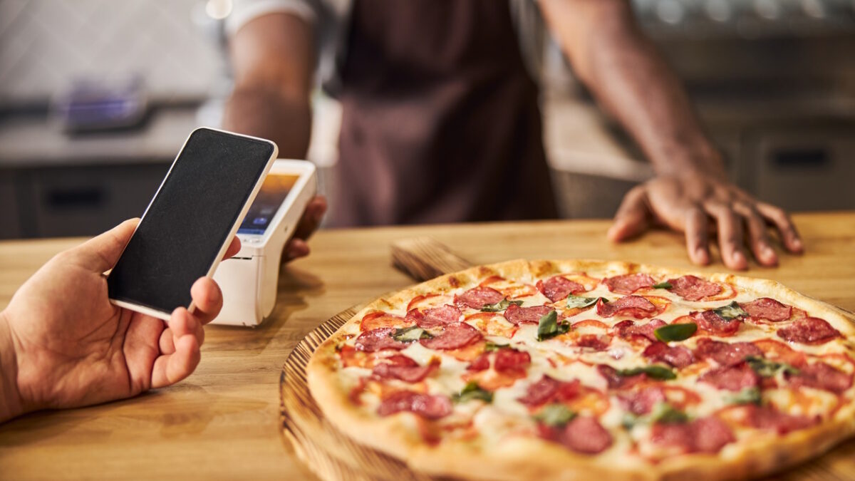 Close up of Afro American man standing at the counter with freshly baked pizza and using terminal for contactless payment while customer holding mobile phone