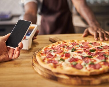 Close up of Afro American man standing at the counter with freshly baked pizza and using terminal for contactless payment while customer holding mobile phone