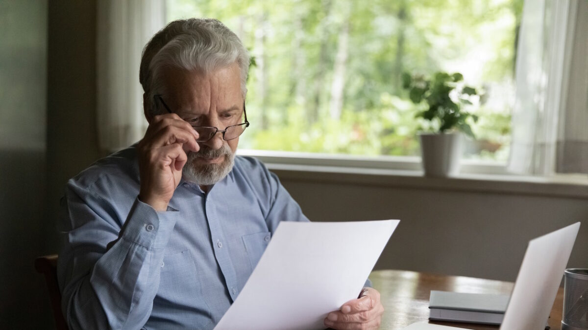 Focused mature 60s - 70s man reading financial document, receives paper bank notification letter at home. Accountant works from home, checking insurance agreement, holds invoice bill for payment