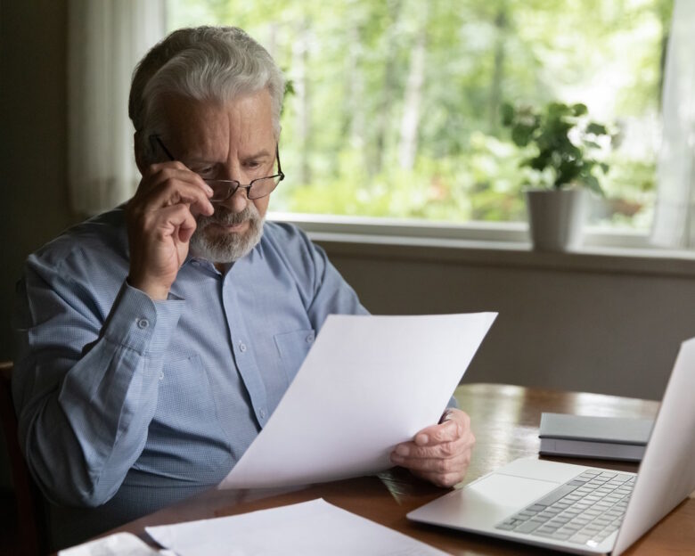 Focused mature 60s - 70s man reading financial document, receives paper bank notification letter at home. Accountant works from home, checking insurance agreement, holds invoice bill for payment