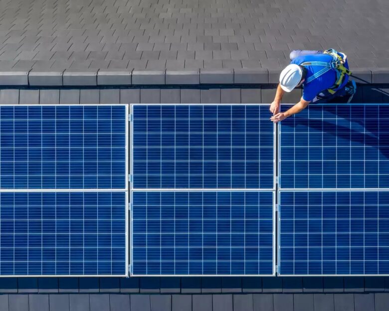 Male engineer in blue suit and protective helmet installing photovoltaic panel system. Professional electrician mounting solar module on roof. Alternative energy ecological concept