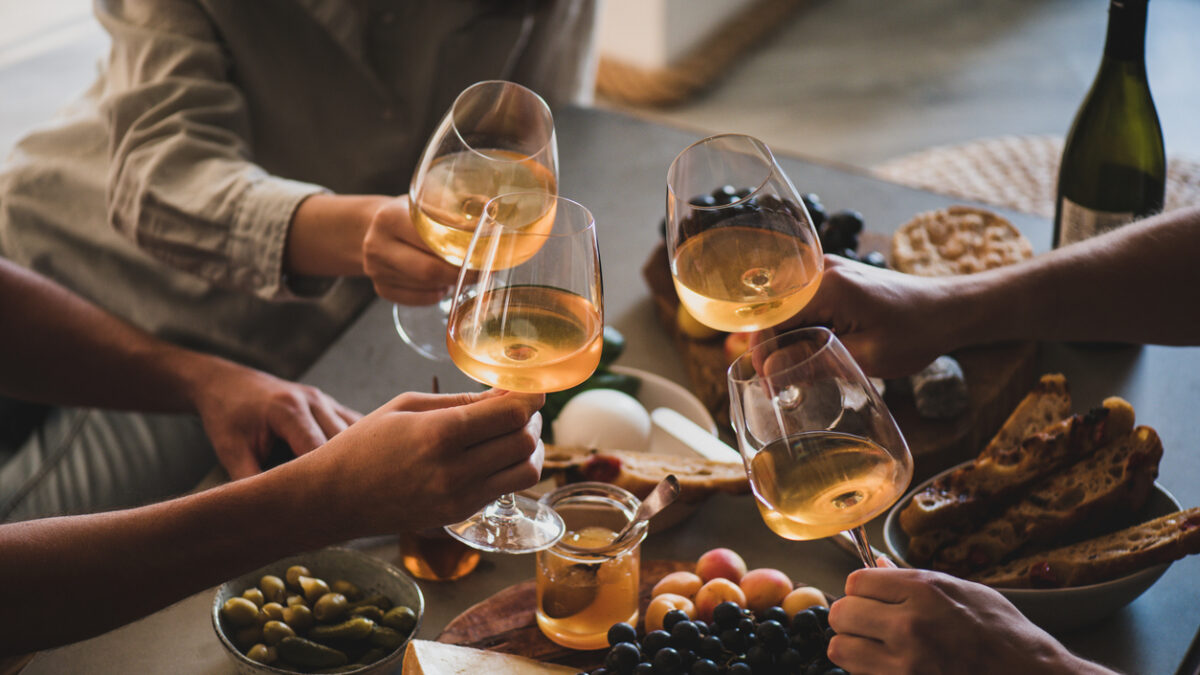 Glasses of white orange or rose wine in hands of celebrating friends over table with various gourmet snacks cheese grape bread. Gathering, celebrating, wine tasting and party concept
