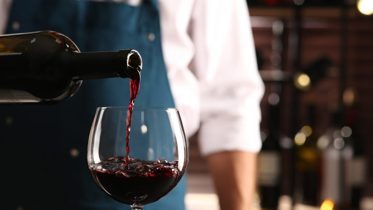 Bartender pouring red wine into glass indoors, closeup. Space for text