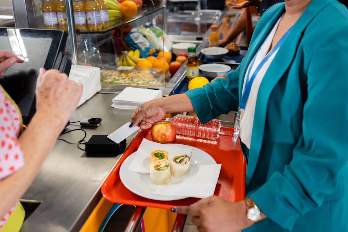 A high angle view of unrecognisable women one of whom is using tap to pay on the card reader. She has some healthy snacks and some water other plate. They are in a school in the North East of England,