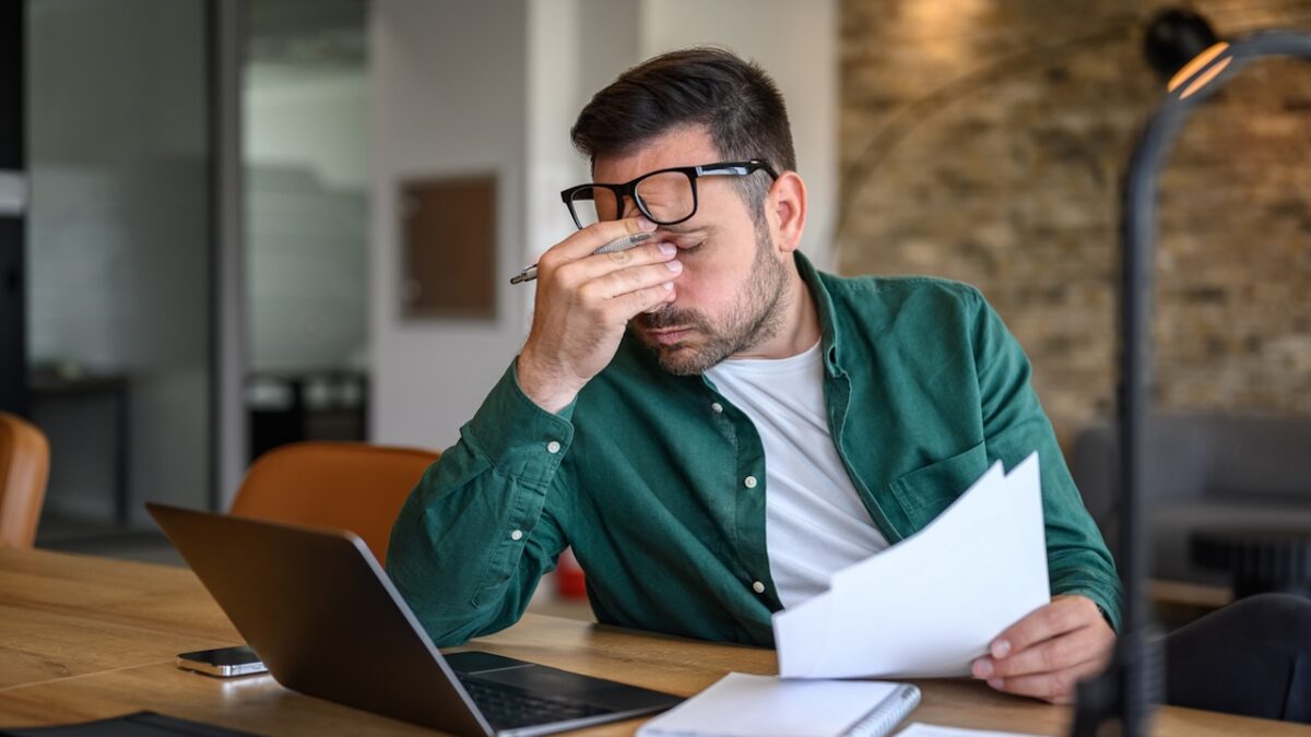 Anxious male financial advisor rubbing eyes while working over laptop and analyzing reports at desk
