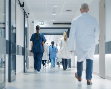 Hospital Hallway with Doctors, Nurses and Specialists in Hospital. Female and Male Physicians, Surgeons, Healthcare Officials Walk Together in Corridor with Their Back to Camera