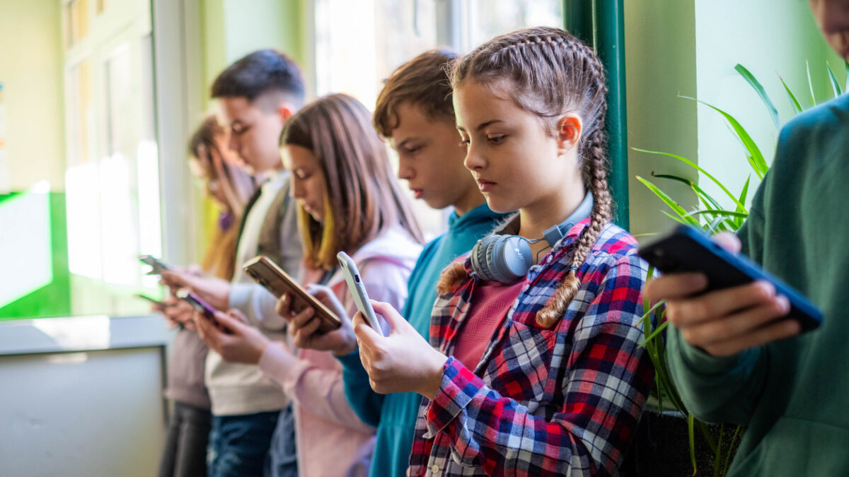 Teenage students are standing in the school hallway, all looking at their phones.