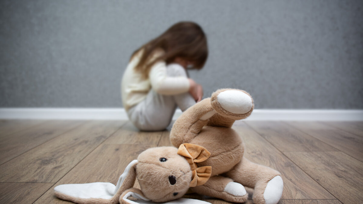A stuffed bunny on the floor, a scared child sitting behind it. Child abuse. Bullying in the family.