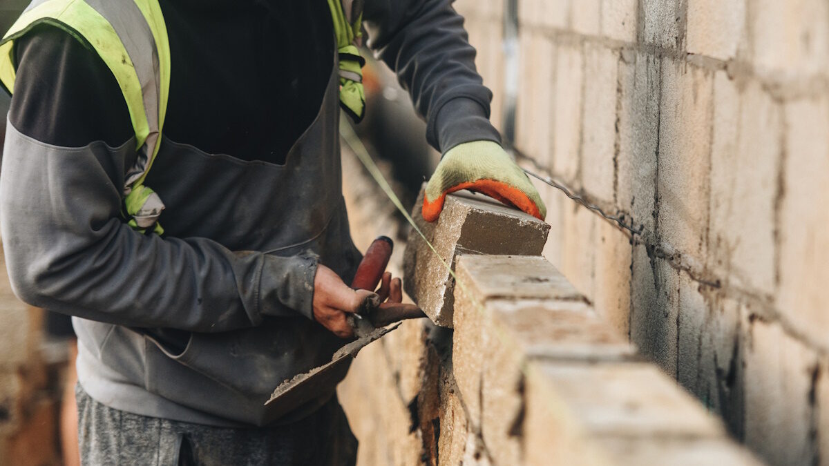 Bricklayer constructing stone wall using stone blocks, cement mortar mix and trowel