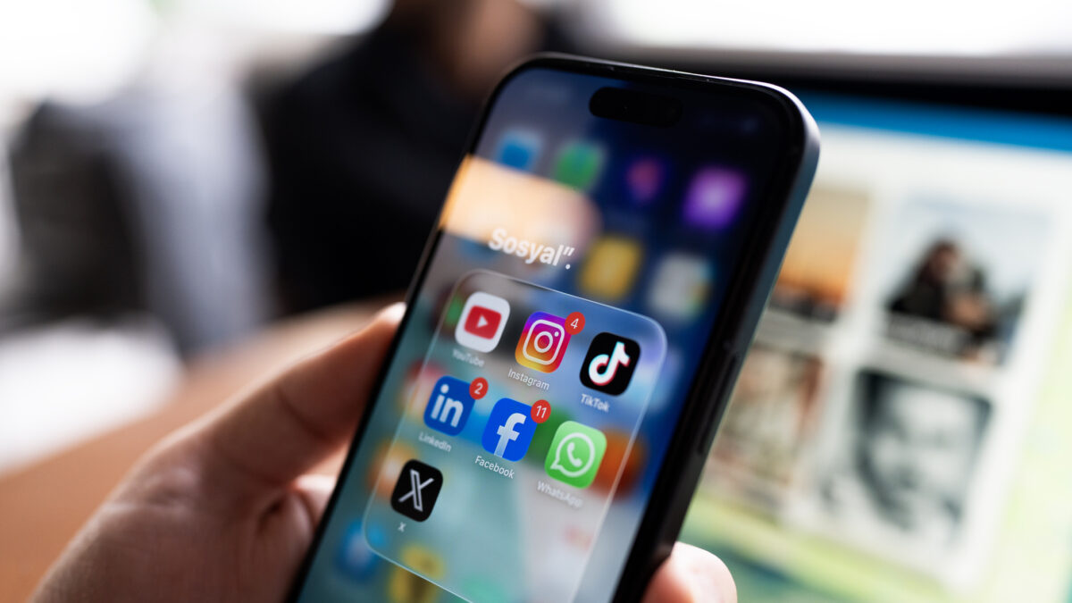Close-up of a person holding a smartphone, displaying popular social media applications including YouTube, Instagram, TikTok, LinkedIn, Facebook, WhatsApp, and X on the screen. A blurred laptop is visible in the background.