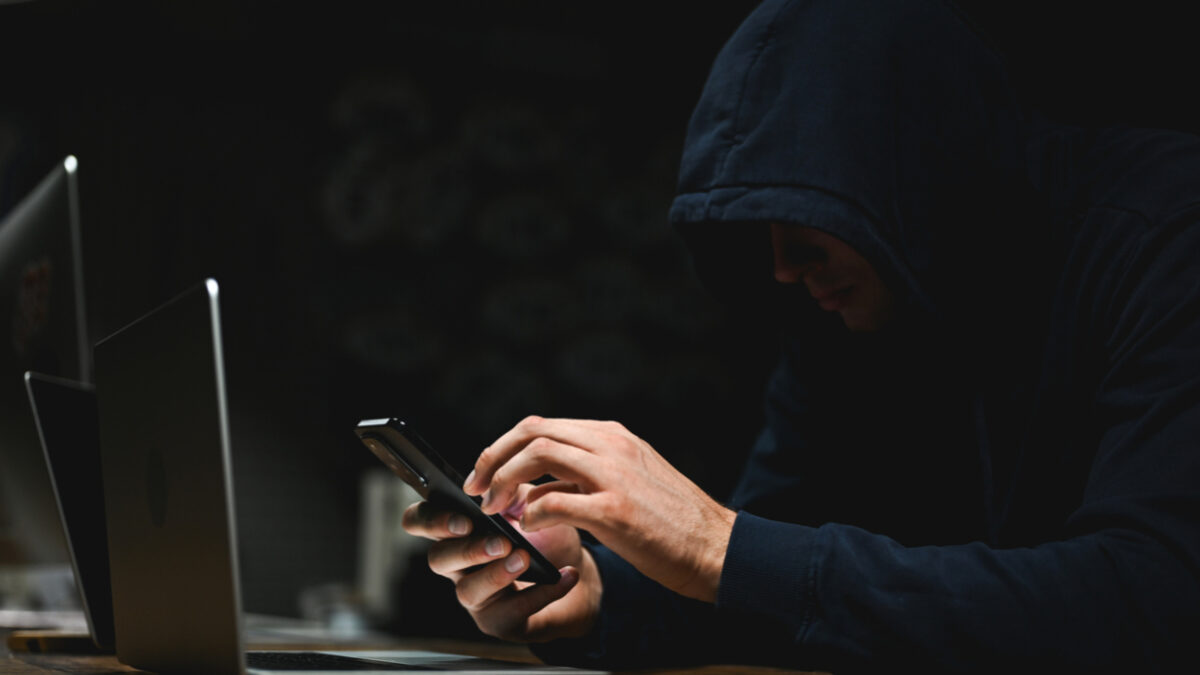 Hooded person using smartphone sitting in front of laptop in dark room