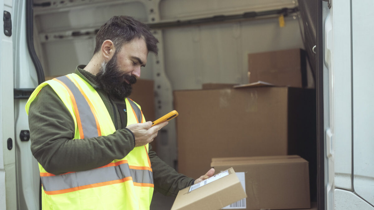 Bearded male courier scanning package label with mobile phone in front of delivery van