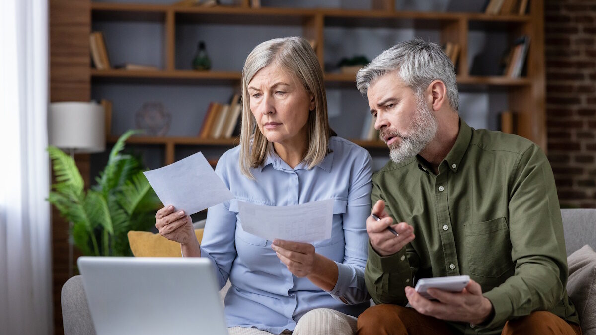 Senior couple working on domestic finances, reviewing documents, using a calculator and laptop to manage household expenses and planning their future financial security in the living room